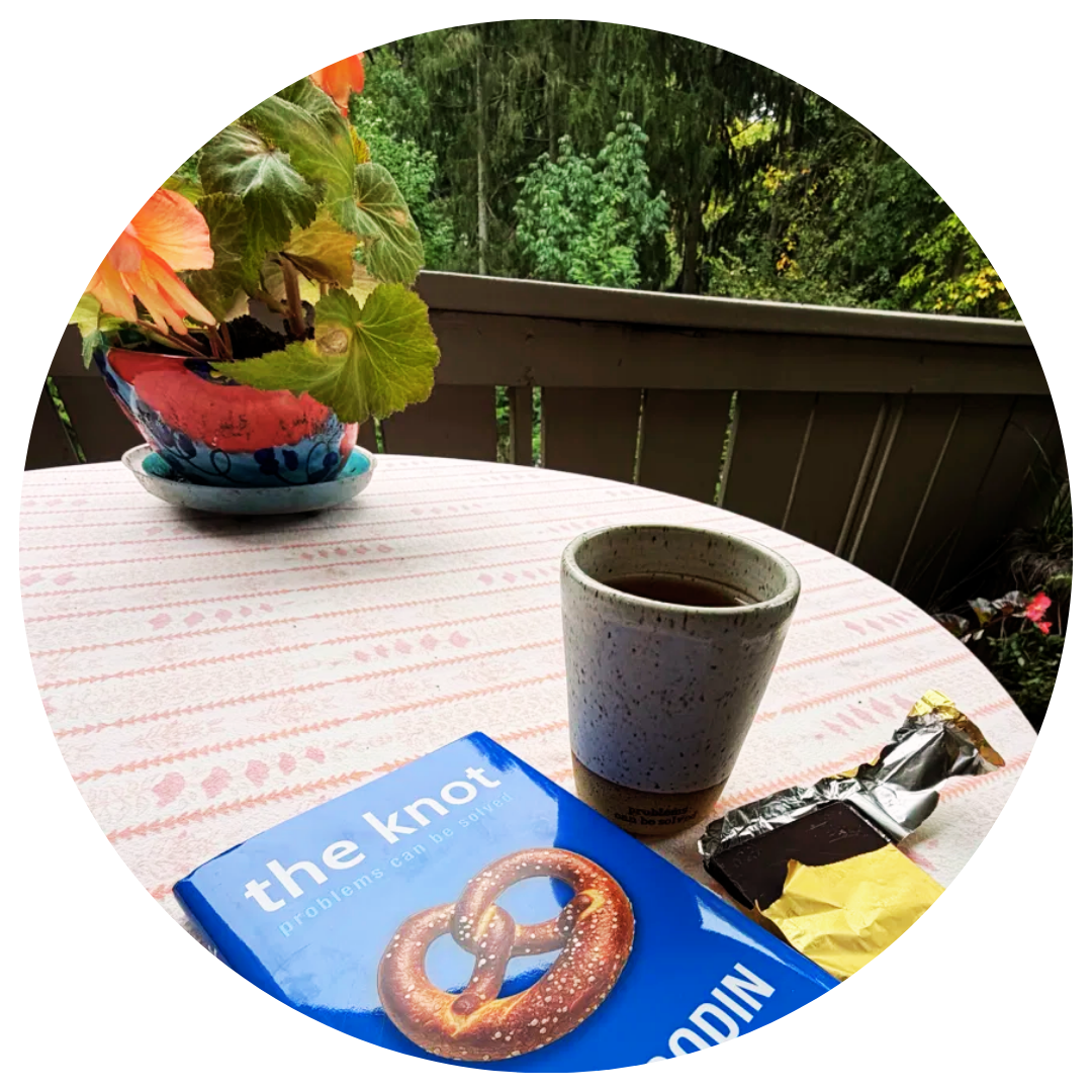 The Knot by Seth Godin with foreword by Mel Robbins, on a garden table with a ceramic mug reading 'problems can be solved,' event swag from Godin's book launch, and a bar of dark chocolate.